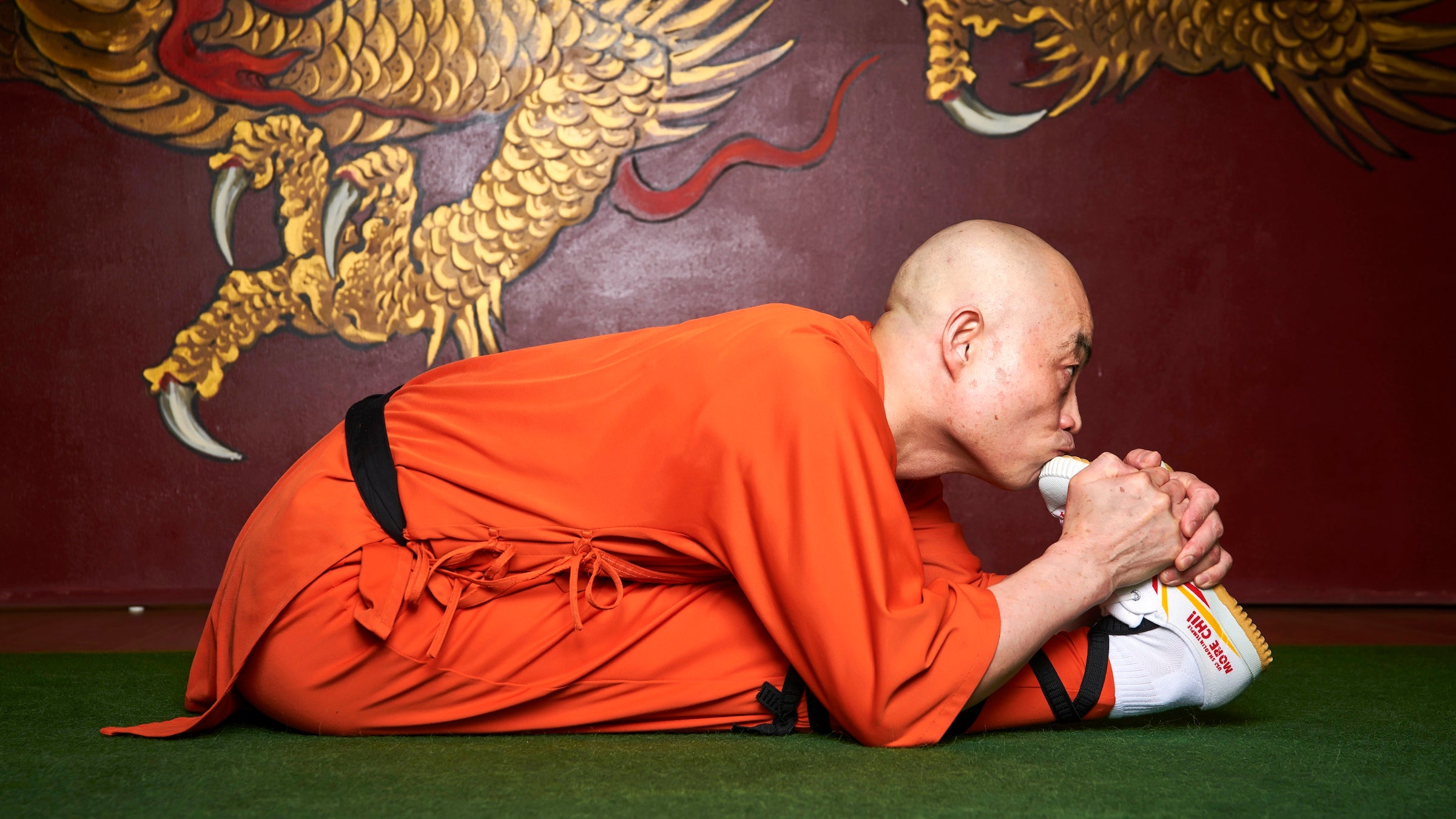Monk in orange robe stretches on a green mat in front of a dragon painting
