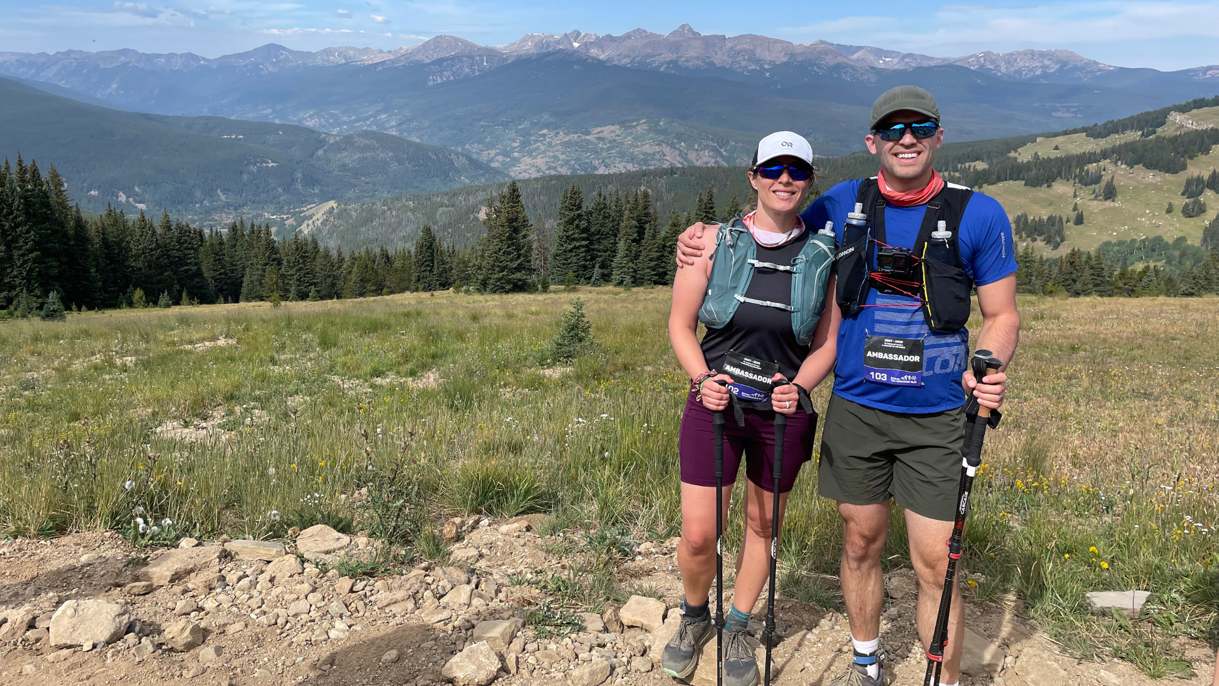 posed photo of husband and wife outside at TransRockies race looking at the camera with trekking poles in hand, mountains in the background